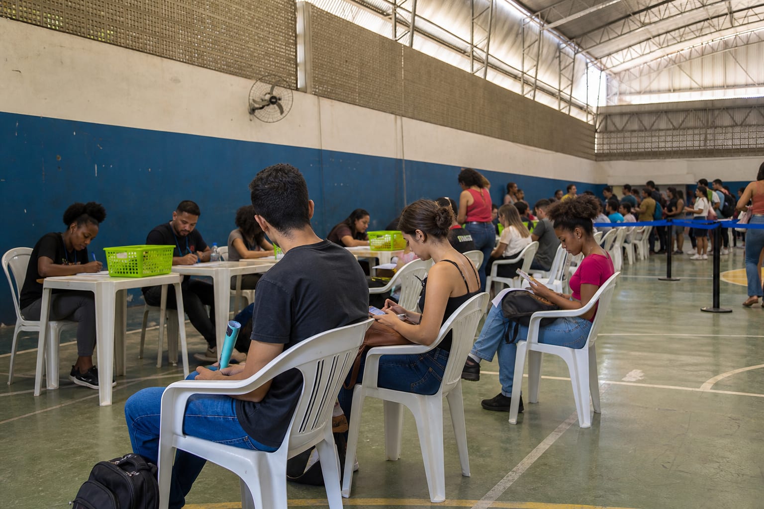 Mutirão de contratação em ginásio de bairro com candidatos sentados em cadeiras de plástico e recrutadores atendendo em mesas ao fundo
